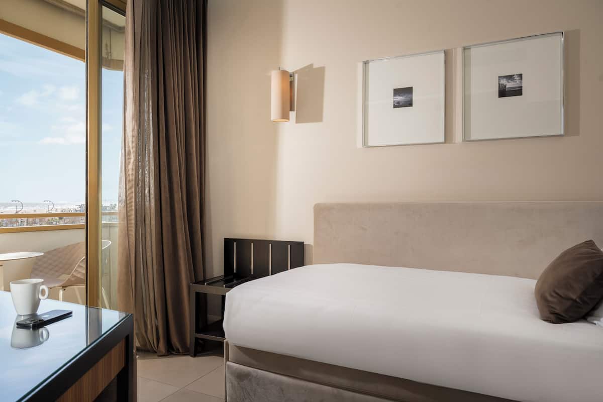 A hotel room with a bed, a desk with a cup and a phone, a chair, a window with curtains, and two framed pictures on the wall.