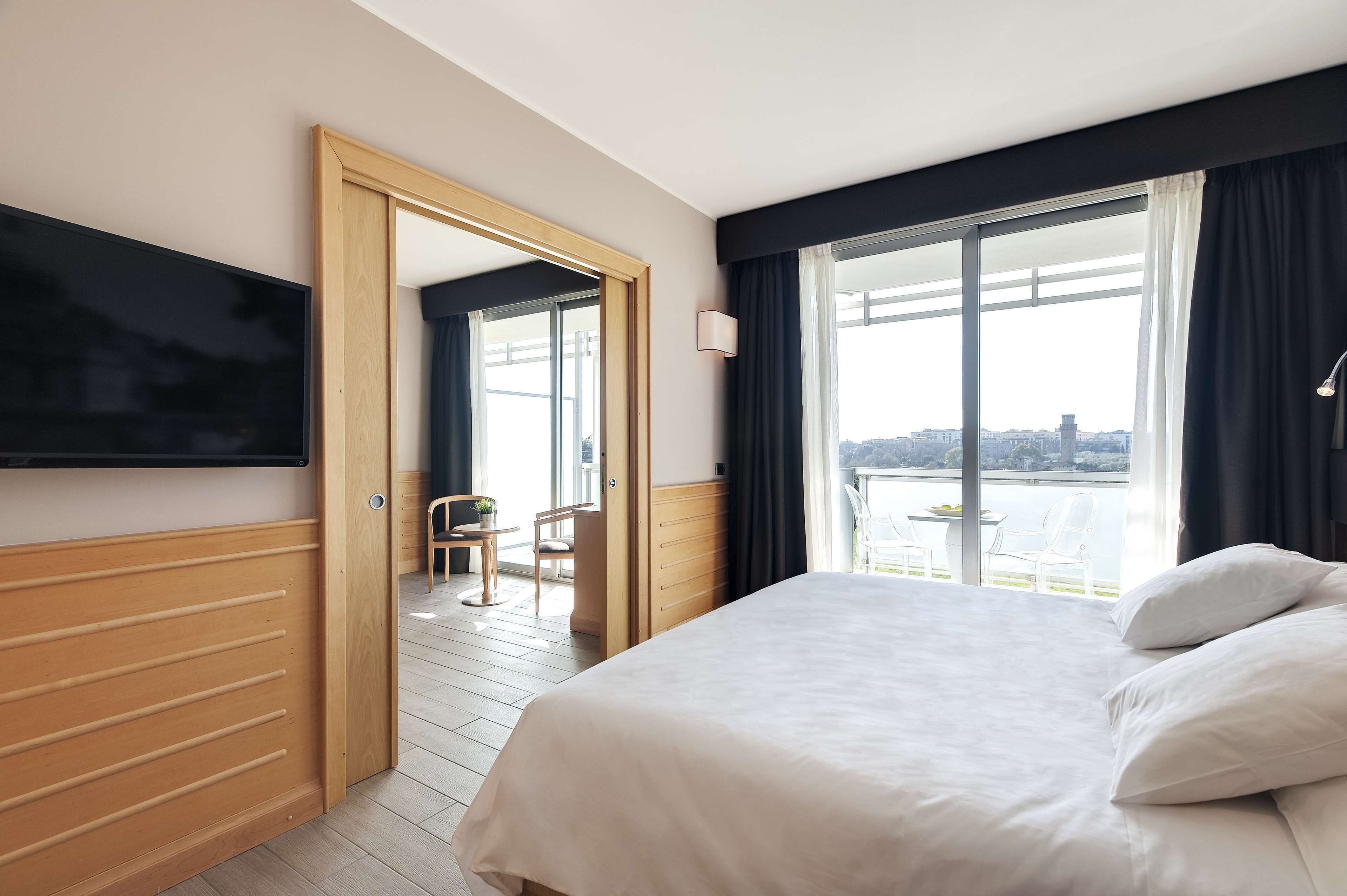A hotel room with a large bed, a TV mounted on the wall, a balcony with a view of the city, and a wooden door leading to a balcony area.
