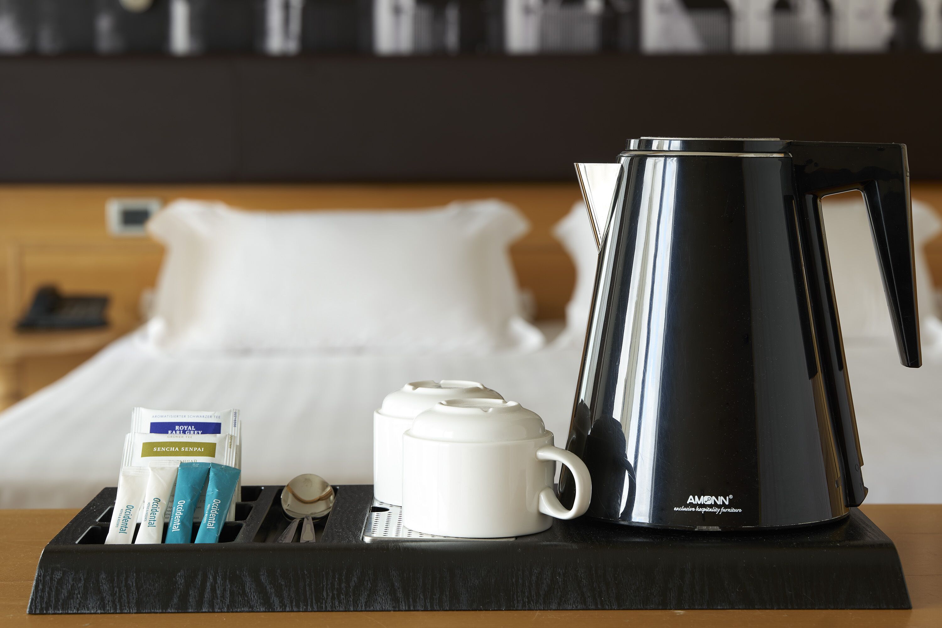 Une chambre d’hôtel avec un lit, une table de chevet sur laquelle se trouve un plateau noir portant une tasse blanche, une bouilloire électrique noire et trois sachets de thé.