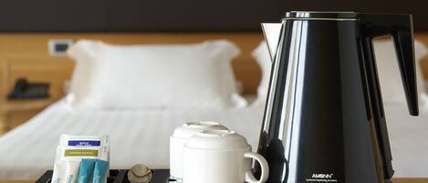 Una camera d'hotel con un letto, un comodino con un vassoio nero che contiene una tazza bianca, un bollitore elettrico nero e tre pacchetti di tè.