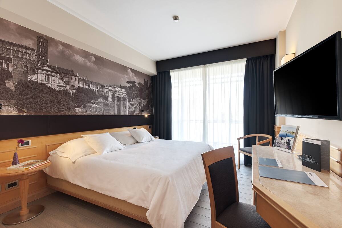 Una habitación de hotel con una cama grande, un escritorio con una silla, un televisor de pantalla plana, una ventana con cortinas y un mural en la pared.