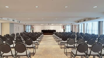 A conference room with rows of chairs facing a podium, large windows with curtains, and a carpeted floor.