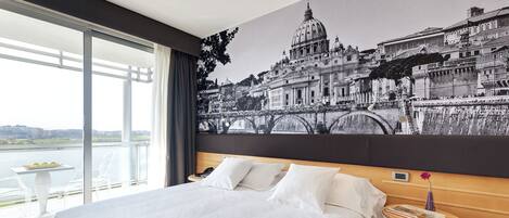 Une chambre d’hôtel avec un grand lit, une table de chevet, une table ornée d’un vase et une vue sur le paysage urbain par la fenêtre.