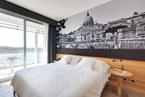 A hotel room with a large bed, a nightstand, a table with a vase, and a view of a cityscape through the window. - Occidental Aran Park (Rome)