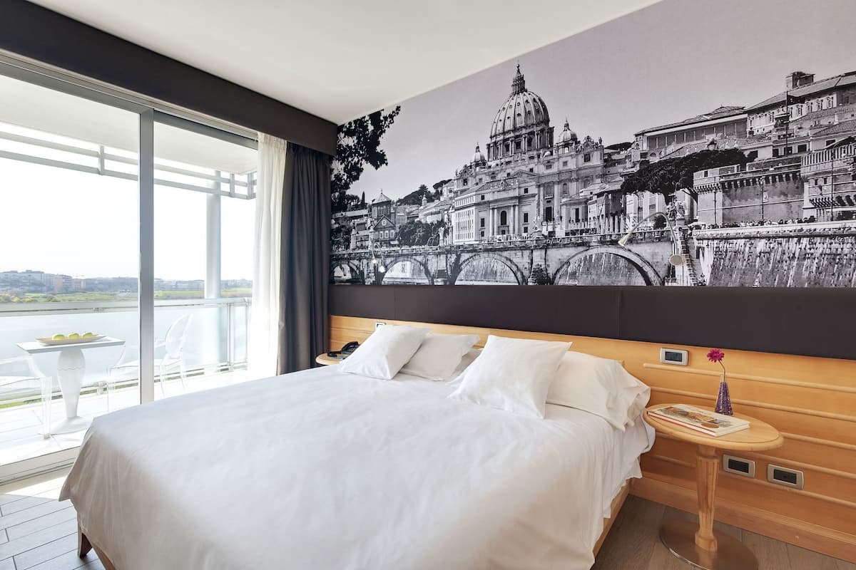 Habitación de hotel con una cama grande, vista a la ciudad, una mesita con un florero y una mesita de noche con un teléfono.