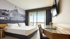 A hotel room with a bed, a desk, a chair, a TV, and a large window with a view. - Occidental Aran Park (Rome)