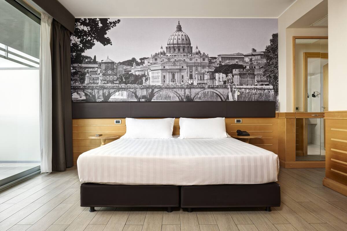 Una habitación de hotel con una cama grande, cabecera de madera y un mural con un paisaje urbano en la pared.