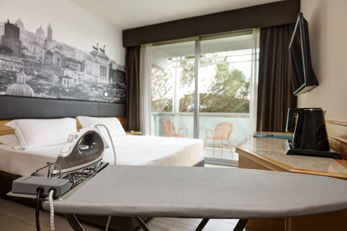 Habitación de hotel con cama, tabla de planchar, televisión de pantalla plana y ventana grande con vista a la ciudad.