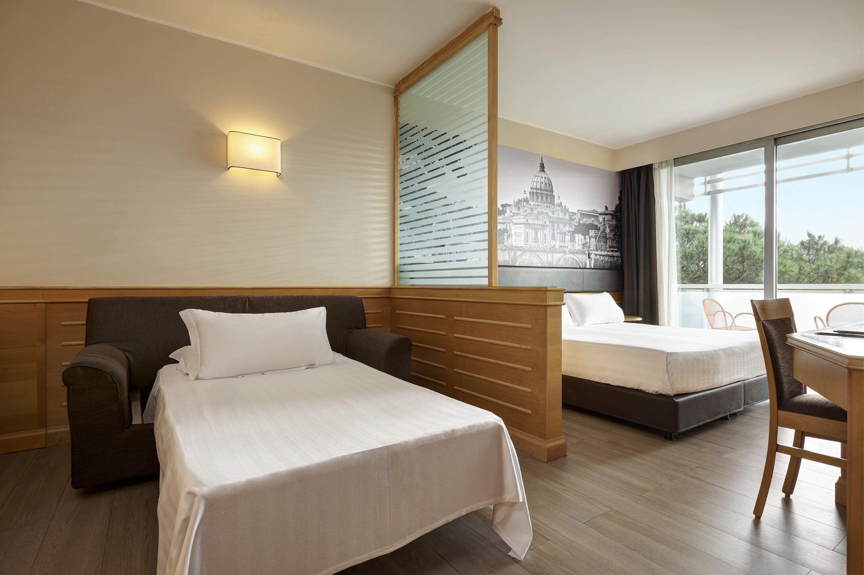 Une chambre d’hôtel avec un grand lit, un plus petit lit, un bureau et une chaise. Il y a une fenêtre donnant sur des arbres et un immeuble.