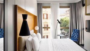 A hotel room with a large bed, a bedside lamp, a chair, and a view of the city through the window. - Le Méridien Visconti Rome (Rome)