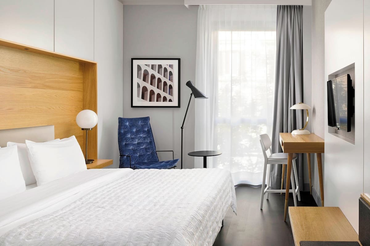 A hotel room with a bed, a desk, a chair, a lamp, a TV, and a window with curtains.