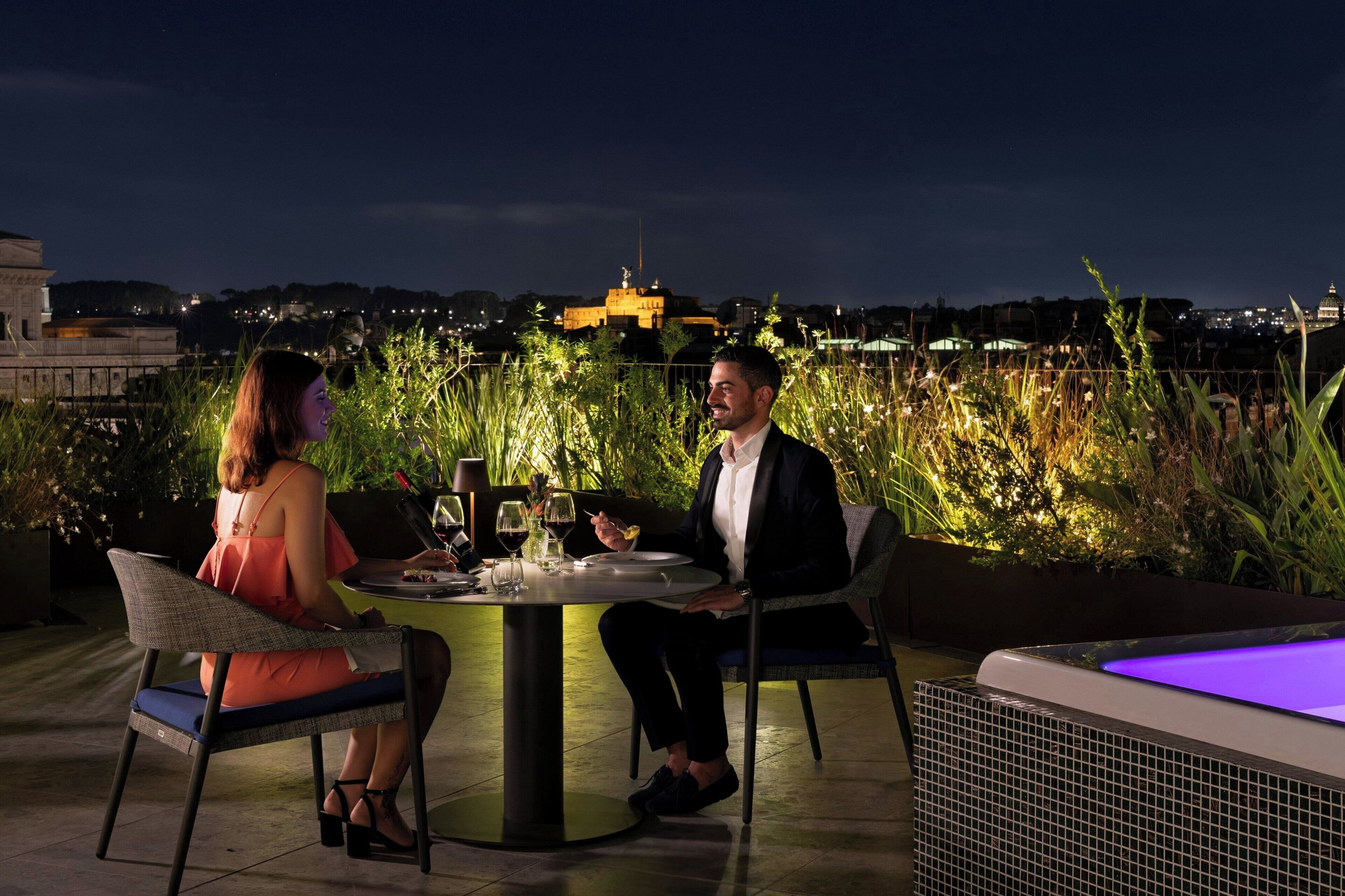 Two people dining at an outdoor table with a cityscape view in the background.