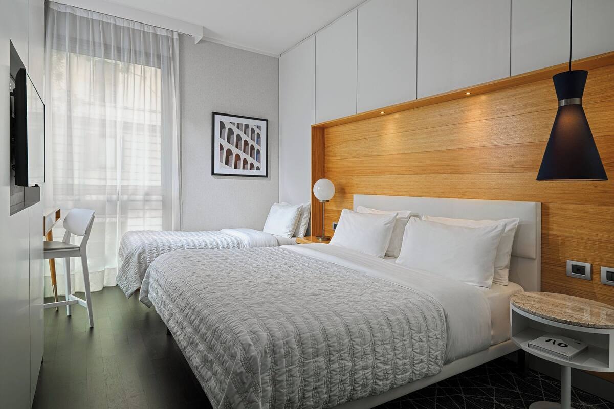 A hotel room with two beds, a wooden headboard, a desk with a lamp, and a framed picture on the wall.