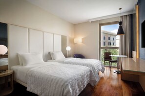 A hotel room with two beds, a desk, a chair, a lamp, and a view of a building through a window.