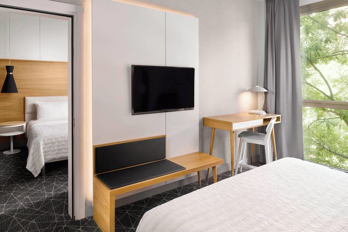 A modern hotel room with a bed, a desk, a chair, a TV, and a window with a view of trees.