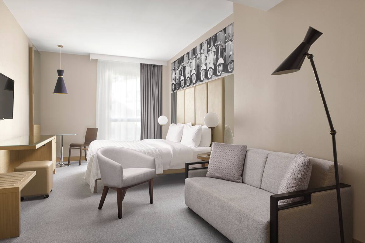 A hotel room with a bed, a sofa, a desk, a chair, a TV, and a lamp.