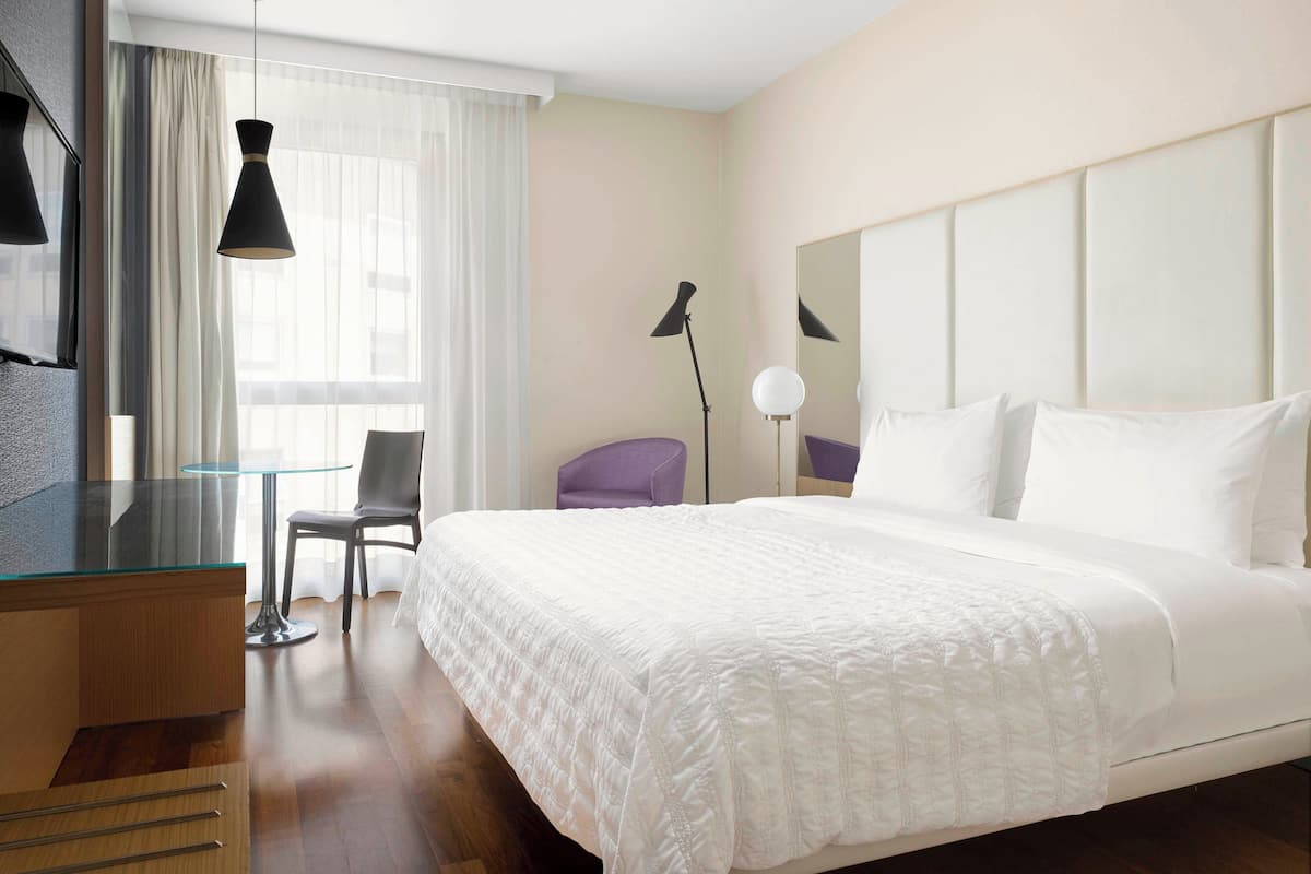 A hotel room with a bed, a desk, a chair, a lamp, and a window with curtains.