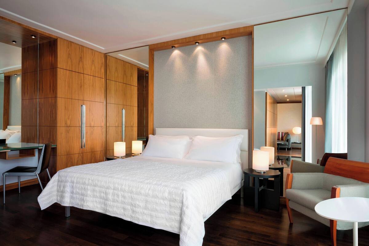 A modern hotel room with a large bed, wooden wardrobe, a seating area with a sofa and armchairs, and a small table.
