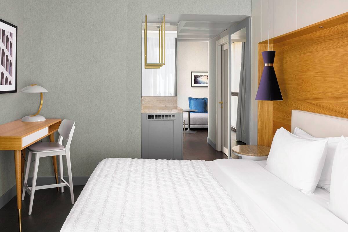 A hotel room with a bed, a desk, a chair, a small table, and a lamp.
