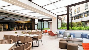 A modern outdoor dining area with wicker chairs and tables, a sofa, and a glass-enclosed space with a view of greenery and buildings. - Le Méridien Visconti Rome (Rome)
