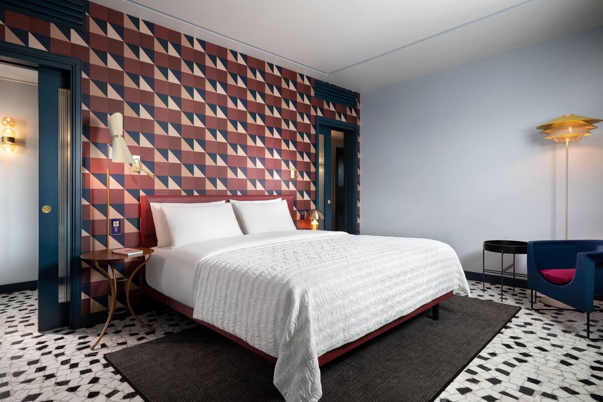 A hotel room with a geometric patterned wall, a neatly made bed, a small side table, a blue chair, and a floor lamp.