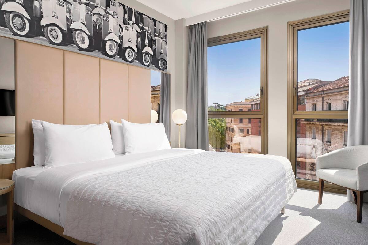 A hotel room with a large bed, a headboard with a cityscape mural, a window with a view of buildings, and a chair.