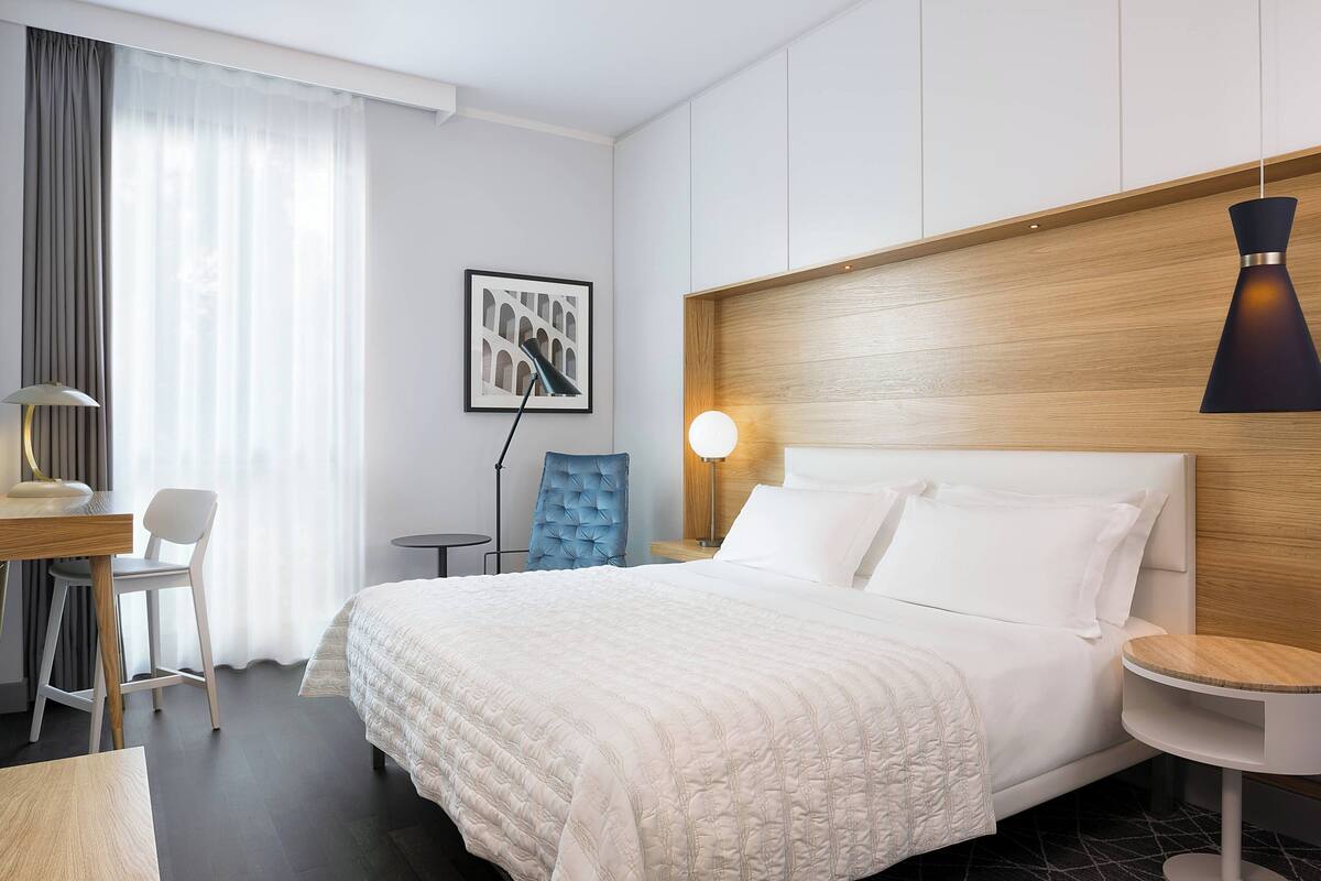 A hotel room with a bed, a desk, a chair, a lamp, and a wall-mounted picture.