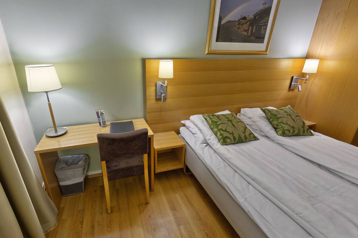A hotel room with a wooden headboard, a desk with a lamp, a chair, and a bed with green pillows.