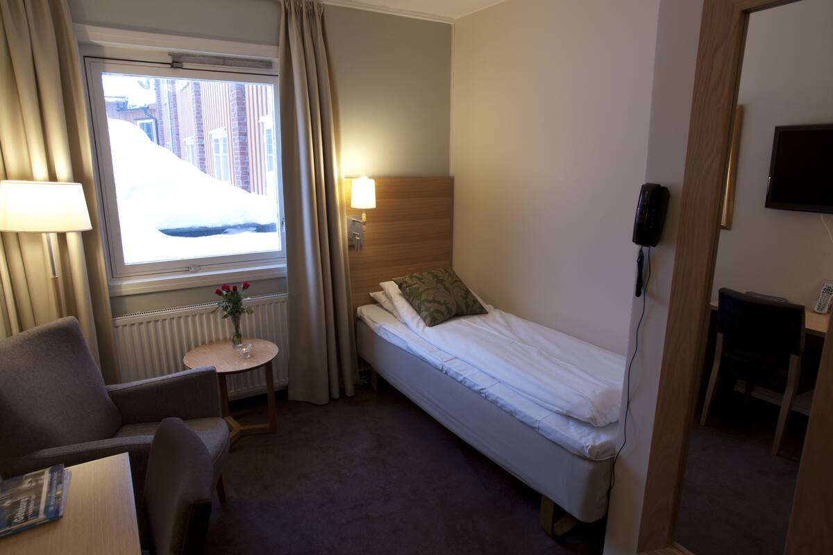 A small hotel room with a single bed, a chair, a small table with a lamp, and a window with curtains.