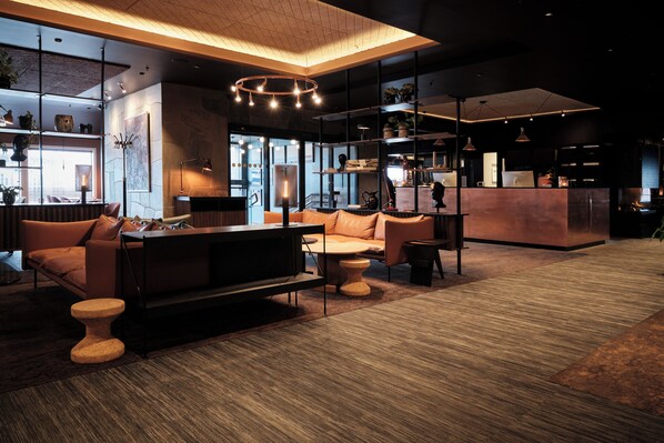 A modern hotel lobby with a seating area, a reception desk, and a kitchen in the background.