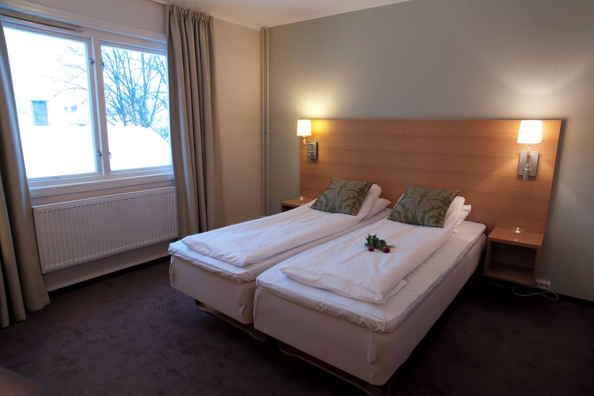 A hotel room with two single beds, a window with curtains, a radiator, and two bedside lamps.