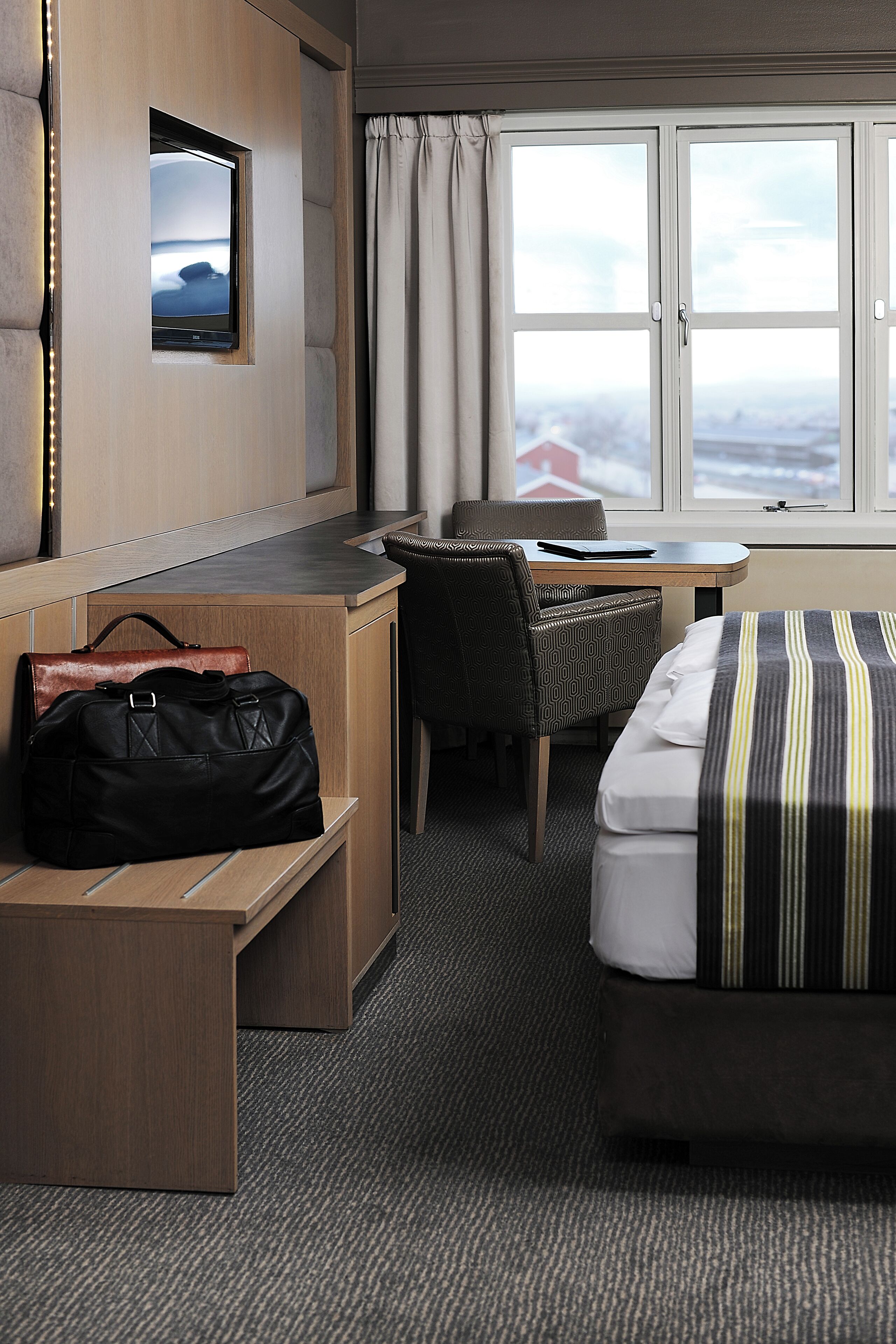 A hotel room with a bed, a desk, a chair, and a small table. There is a window with curtains, a wall-mounted TV, and a bag on the desk.
