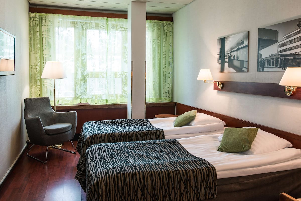 Habitación de hotel con dos camas, una silla y un ventanal con cortinas.