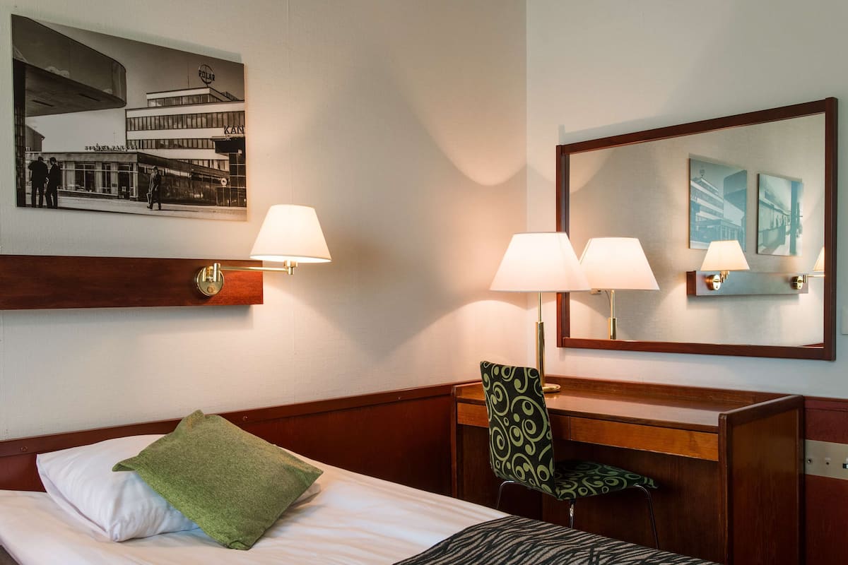 Habitación de hotel con cama, dos lámparas de pared, un escritorio con silla, un espejo y un cuadro en la pared.