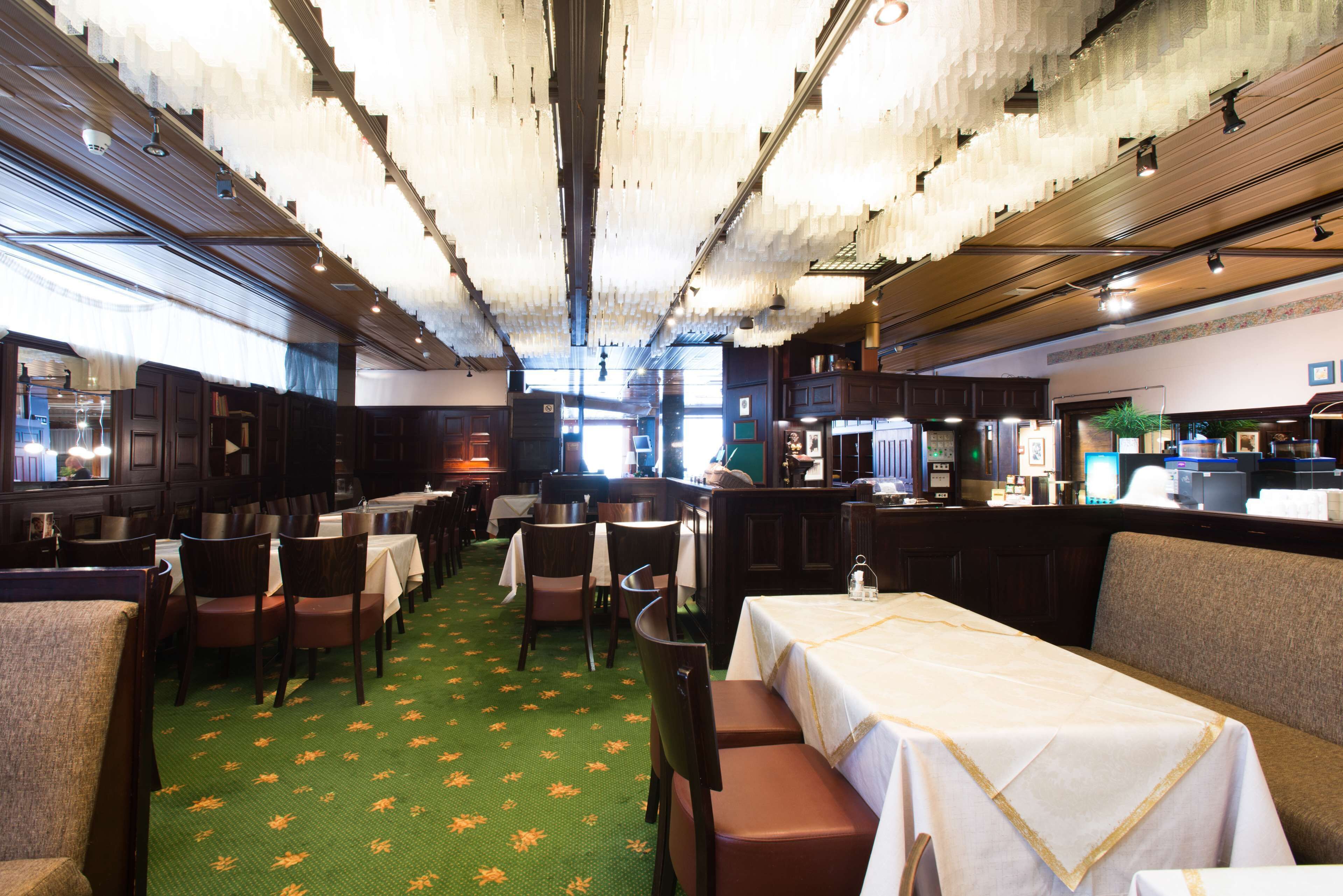 Un restaurante con cabinas y mesas, alfombra verde con diseños anaranjados, y un techo alto con luces colgantes.