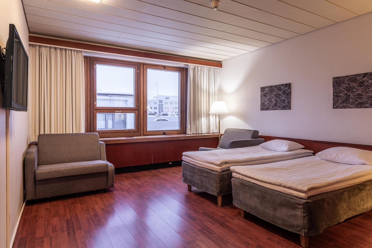 Habitación de hotel con piso de madera, dos camas individuales, un sofá, un televisor y una ventana con cortinas.