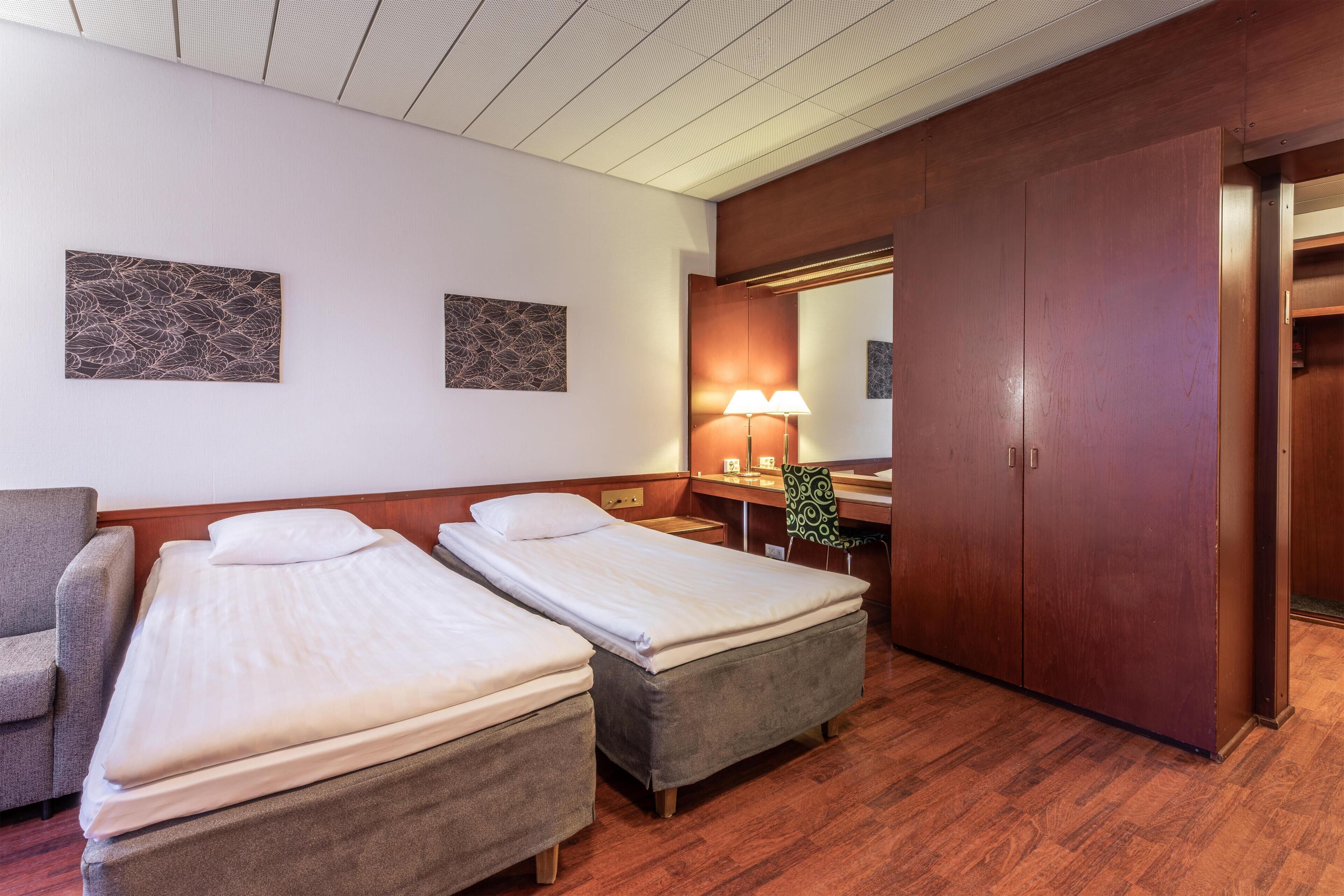 A hotel room with two single beds, a desk with a chair, a lamp, and a wardrobe.