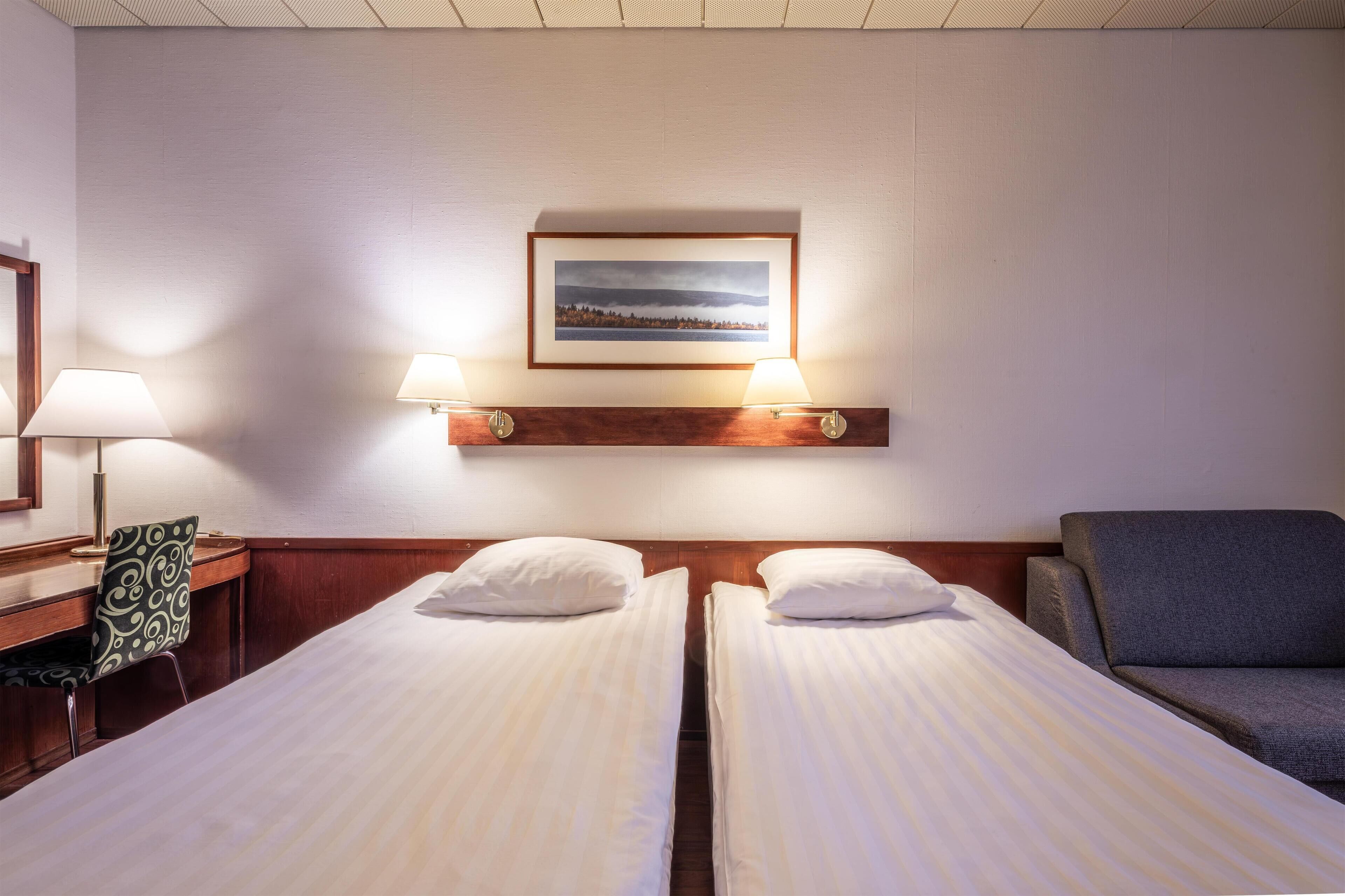 A hotel room with two single beds, a desk with a chair, a sofa, and a framed picture on the wall.