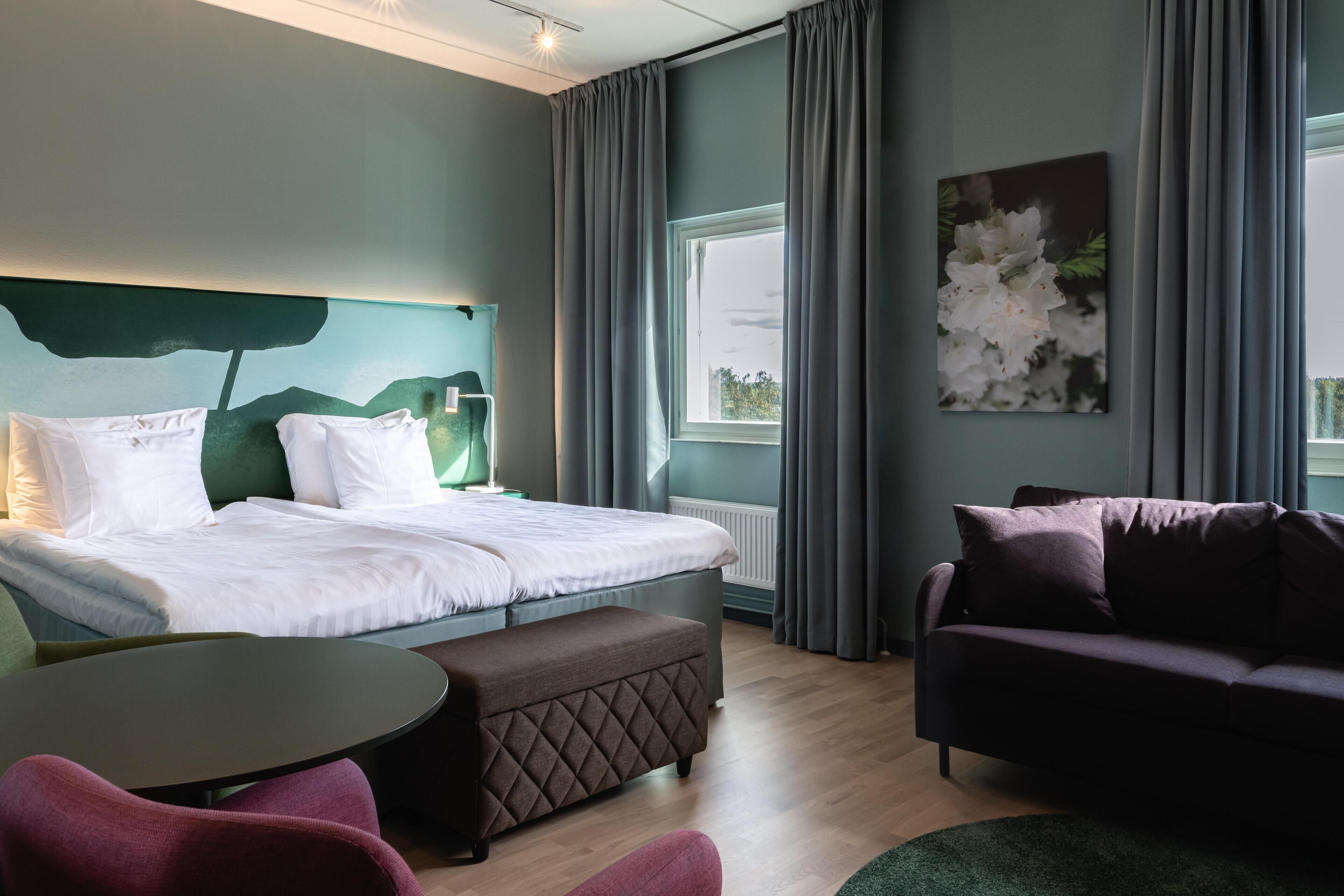 A modern hotel room with a neatly made bed, a sofa, a small table, and a large window with curtains.