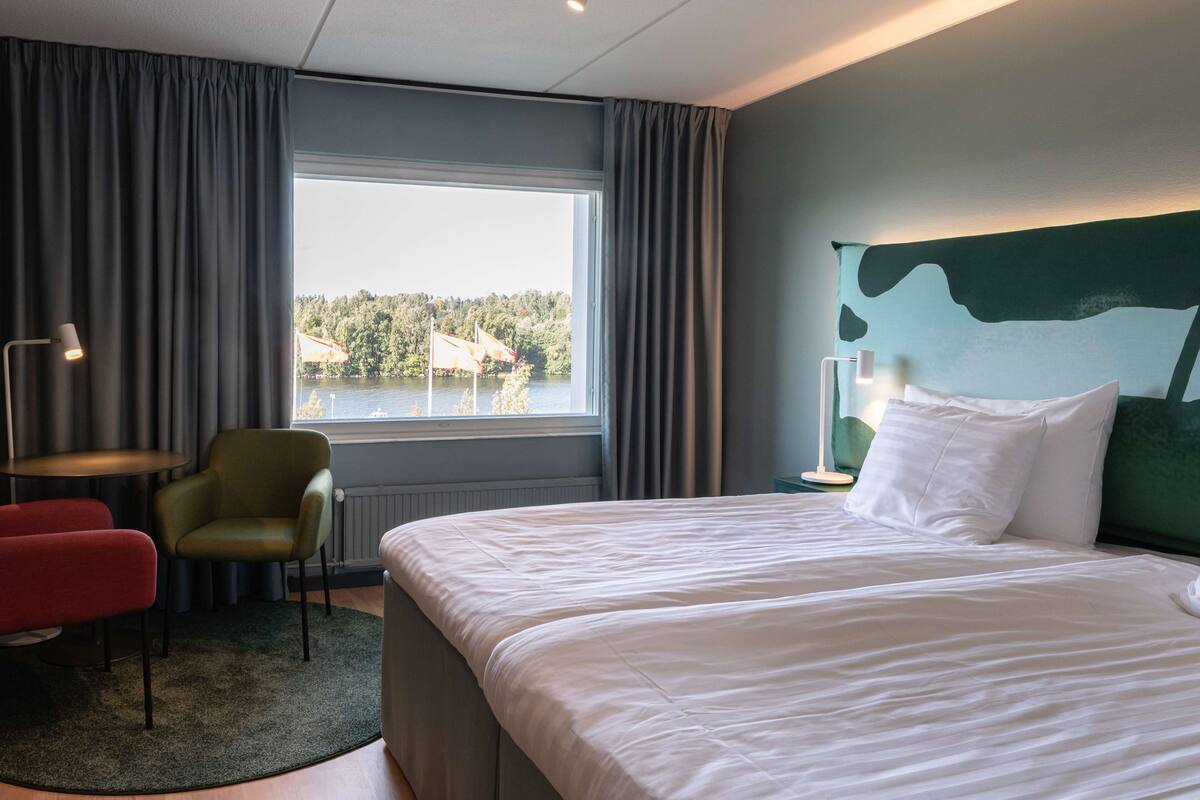 Hotellihuone, jossa on suuri sänky, vihreä nojatuoli, punainen tuoli ja ikkunasta avautuva näkymä järvelle.