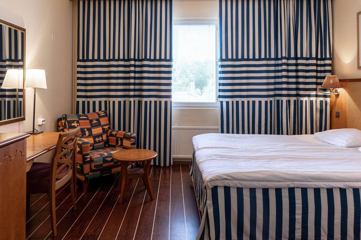 Hotellihuone, jossa on raidalliset verhot, sänky, tuoli, pöytä ja lamppu.