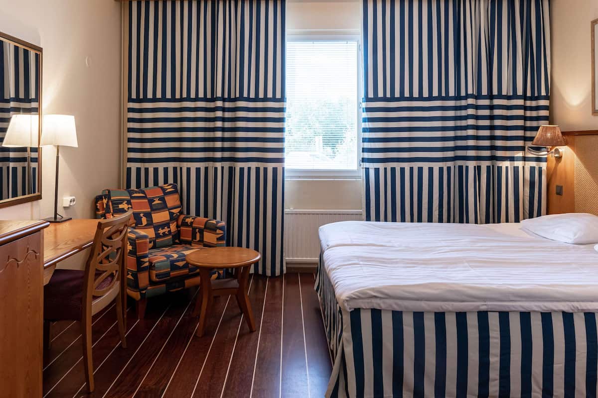Quarto de hotel com cama, uma cadeira, uma mesinha, luminária e cortinas listradas.