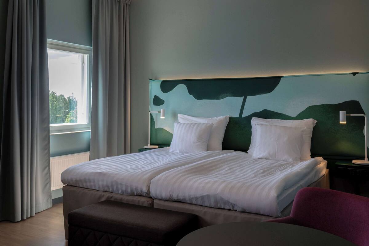 Hotellihuone, jossa on suuri sänky, kaksi yöpöytävalaisinta, yöpöytä, ikkuna verhoilla ja näkymä vehreään maisemaan.