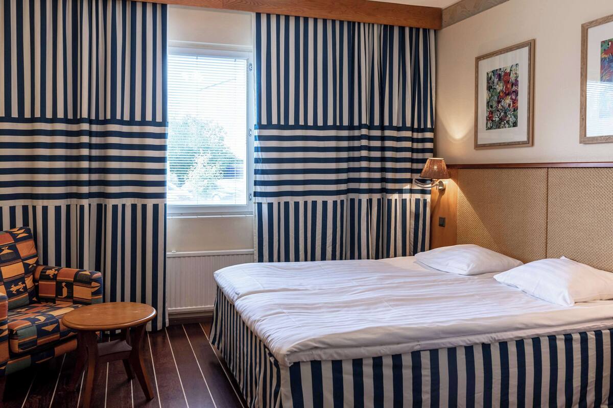 Hotellihuone, jossa on raidallinen päiväpeite, puinen pääty, pieni pyöreä pöytä, kuviollinen nojatuoli ja ikkuna, jossa on verhot.