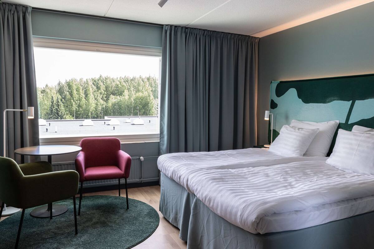 Hotellihuone, jossa on sänky, työpöytä, tuoli ja ikkunasta avautuva näkymä puita kohti.