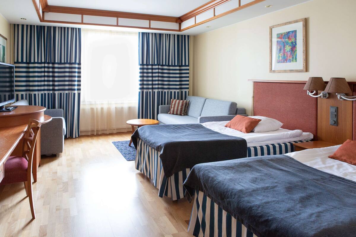 Hotellihuone, jossa on kaksi sänkyä, työpöytä, tuoli, televisio ja ikkuna, jossa on verhot.