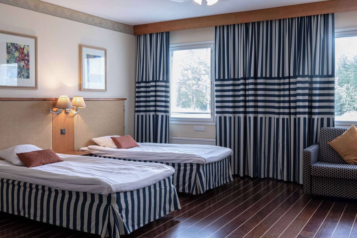 Hotellihuone, jossa on kaksi sänkyä, sohva ja ikkuna, jossa on raidalliset verhot.