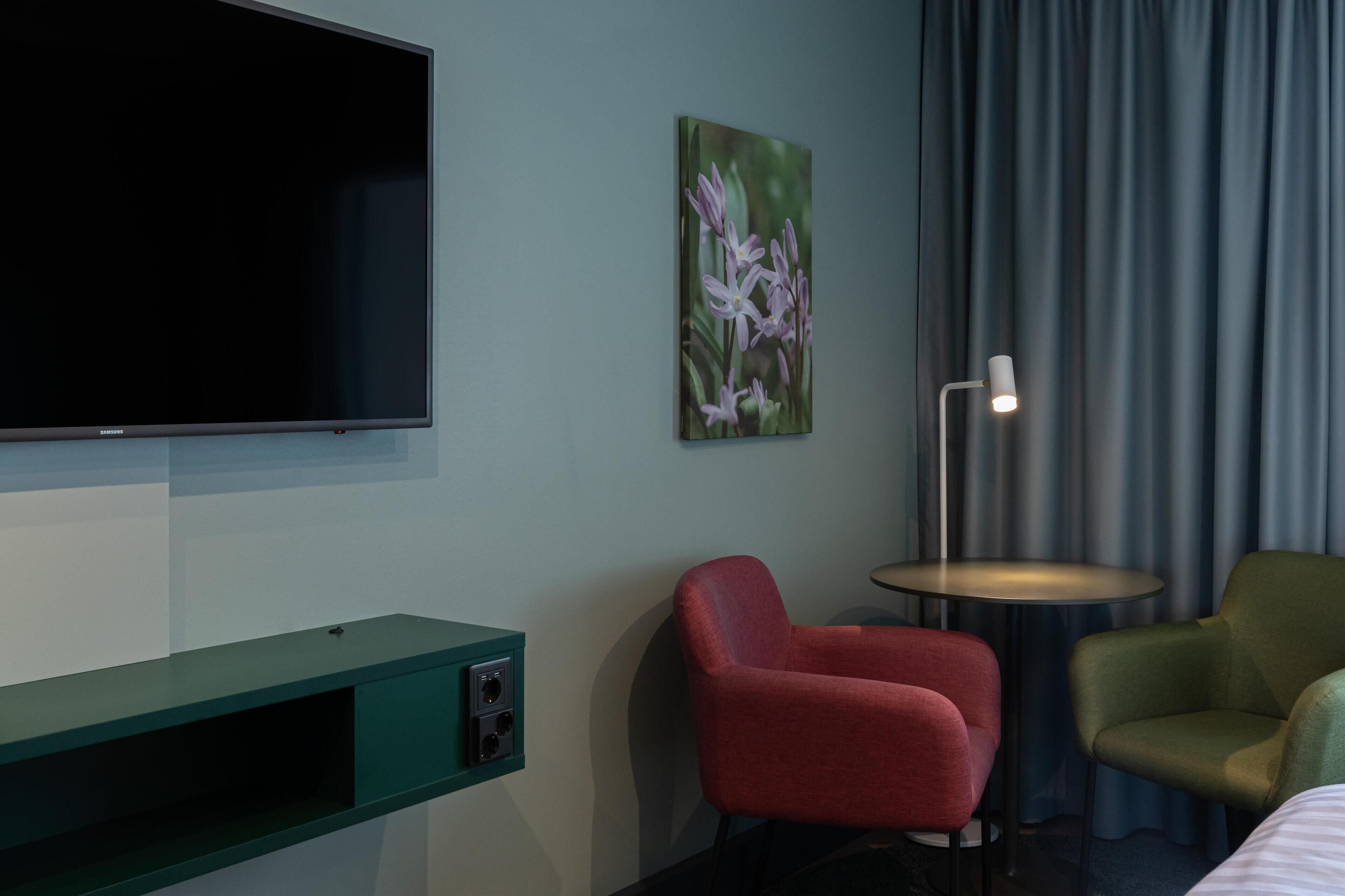 A hotel room with a flat-screen TV, a painting of flowers, a red armchair, a green armchair, and a small round table with a lamp.