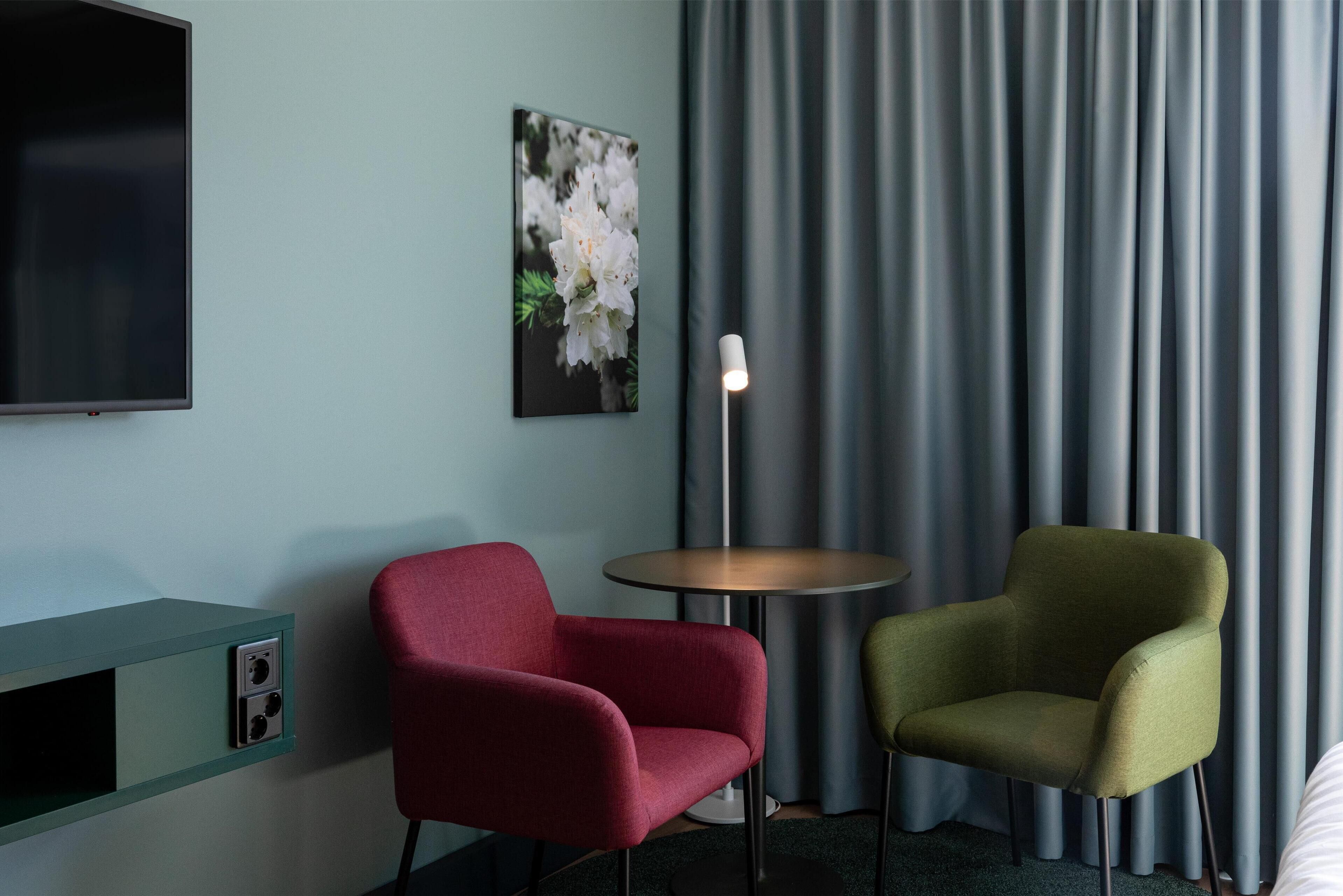 A hotel room with a flat-screen TV, a painting of flowers, two armchairs, a small round table, and a lamp.