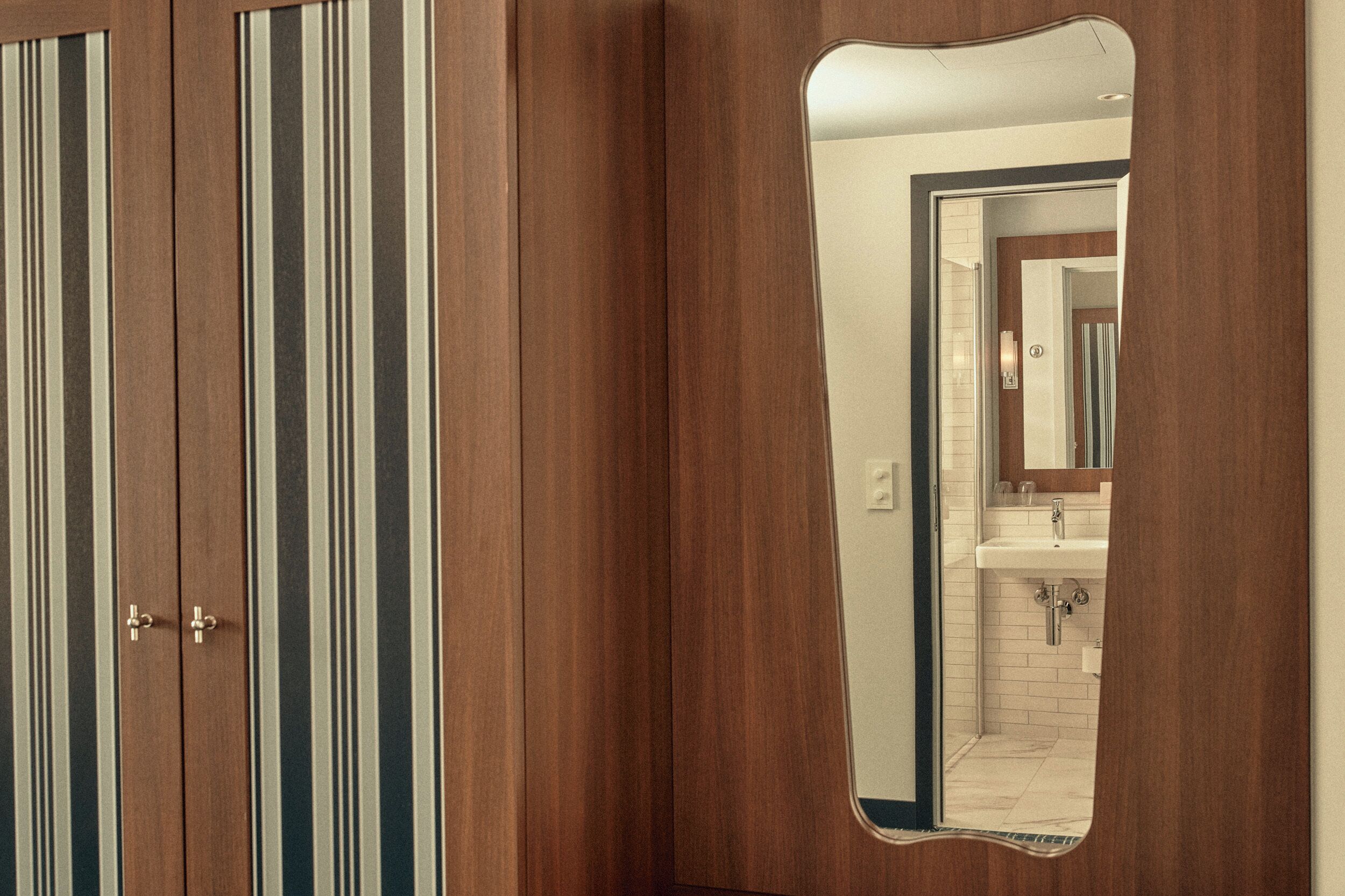 Une armoire en bois à rayures verticales, dotée d’un miroir à cadre ondulé qui reflète une salle de bain avec un lavabo et un miroir.